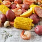 cooked corn on the cob, shrimp, red potatoes, and sausage in a pile on a newspaper with lemon wedges.