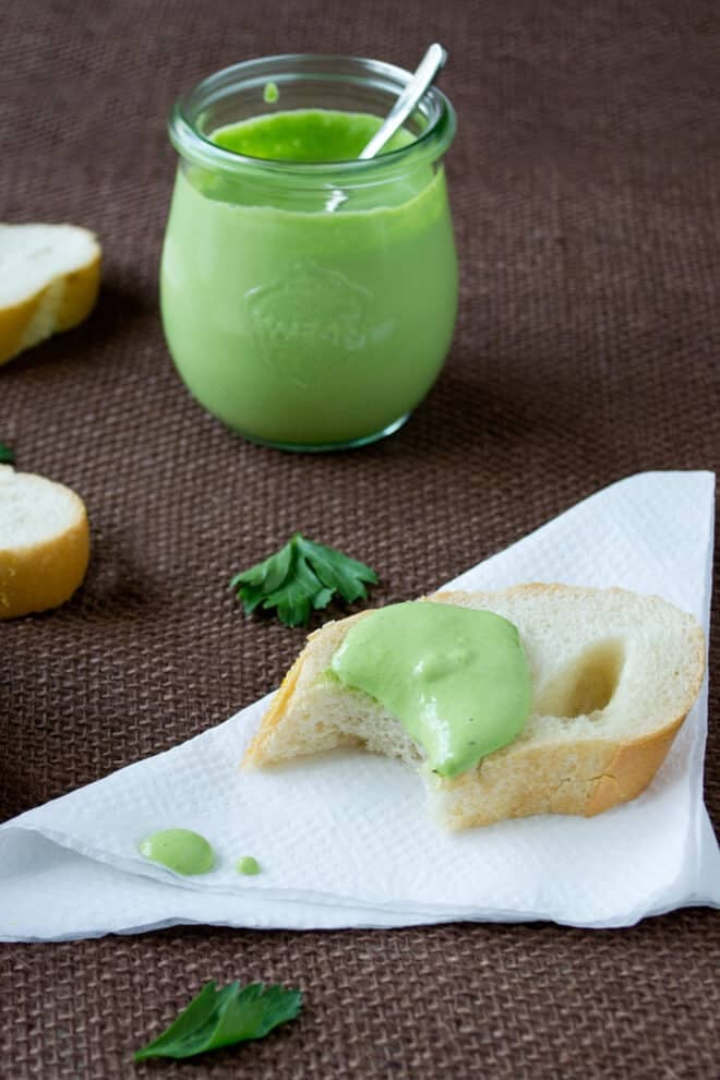A slice of baguette with green milk mayonnaise on it, on a white napkin. A jar of more mayonnaise and more slices of baguette are in the background.