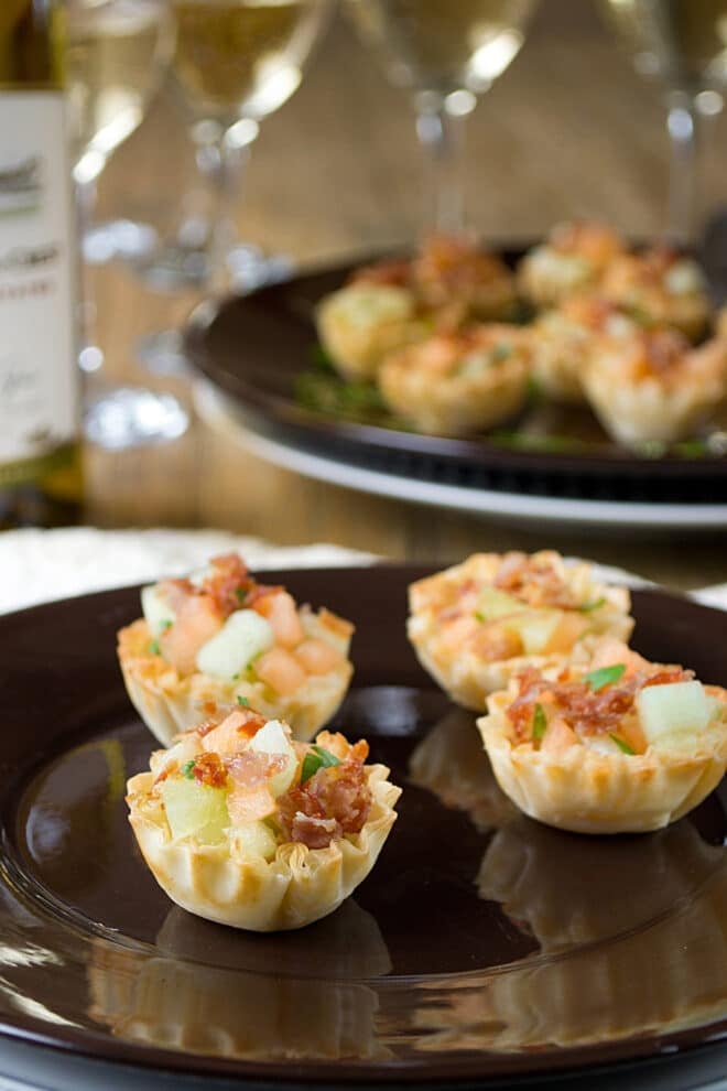 Small phyllo cups filled with small cubes of cantaloupe, honey dew melon, and crispy prosciutto. Four of the cups are arranged on a plate, with a plate of more in the background along with glasses of white wine.