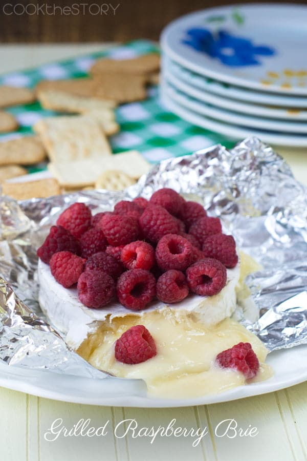 Grilled Brie with fresh raspberries on top, all on tin foil on a plate, crackers in background.
