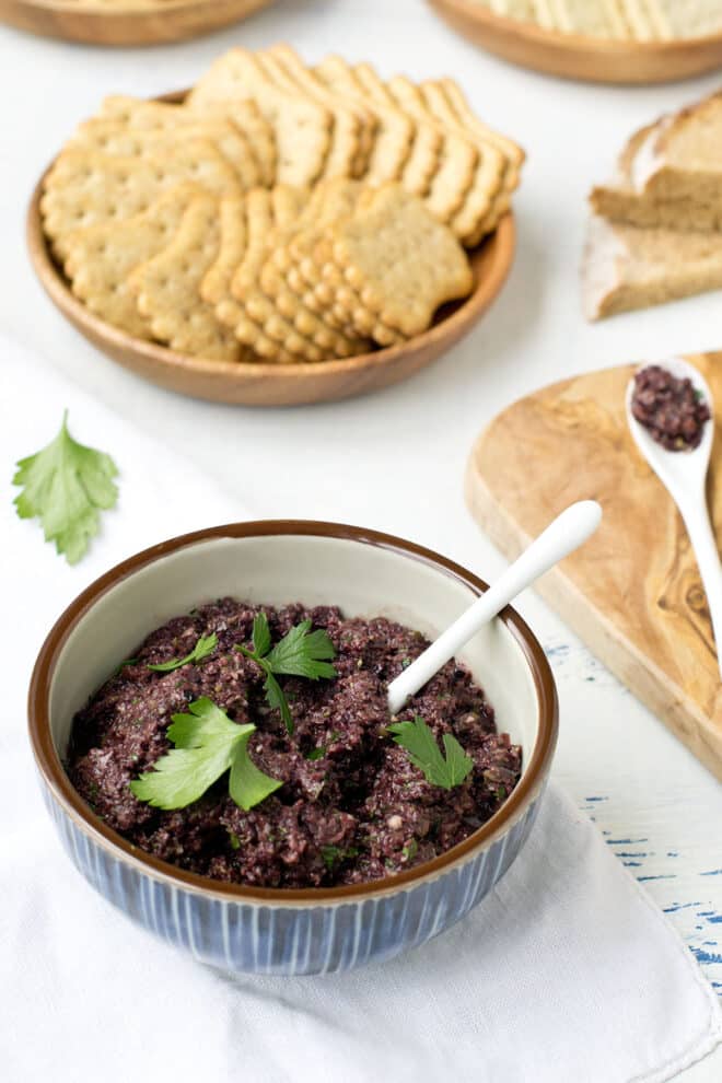 A blue bowl full of dark-colored olive tapenade, garnish with fresh Italian parsley leaves, crackers in the background