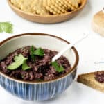 A bowl full of tapenade made from kalamata olives, garnished with fresh parsley leaves, crackers in the background