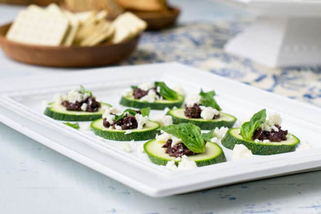 Slices of raw zucchini topped with tapenade, feta, and fresh basil leave arranged on a plate.