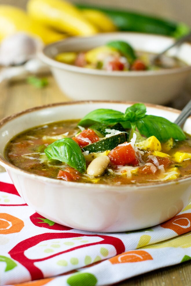A bowl of minestrone soup. Tomatoes and summer squash are visible at the top along with a garnish of basil. Another bowl of soup and some whole zucchini and summer squash are in the background.