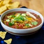 Bowl of chicken tortilla soup topped with cheese, green onion, and diced avocado. Surrounded by corn tortilla chips.