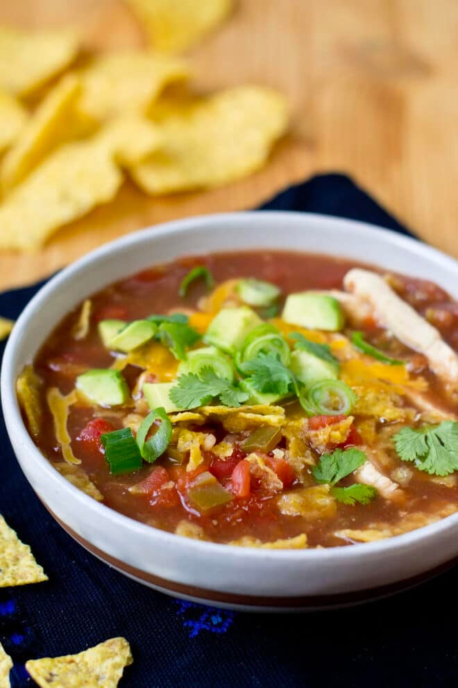 Bowl of chicken tortilla soup topped with cheese, green onion, and diced avocado. Surrounded by corn tortilla chips.
