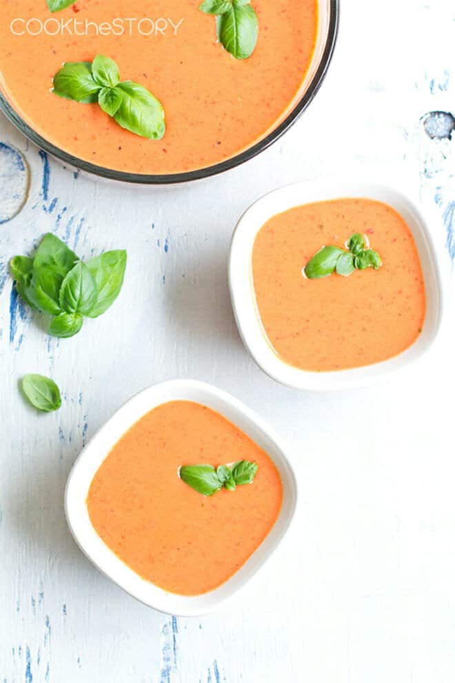White bowls of red pepper soup with fresh basil leaves.
