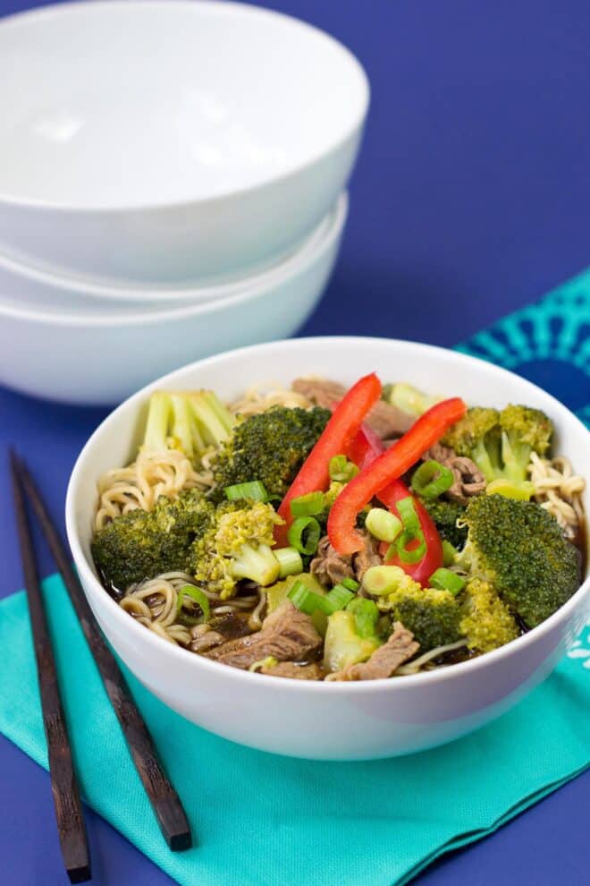 A white bowl with ramen noodles, broth, broccoli, red bell peppers, and green onion. Chop sticks on the side, and a stack of empty bowls are behind.