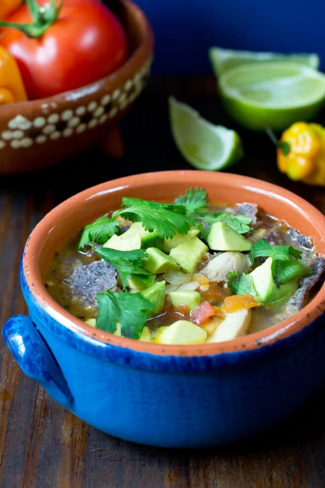A terracotta bowl painted blue on the outside full of soup topped with diced avocado, fresh cilantro, and corn chips. There are fresh ingredients like tomatoes, lime, and habanero pepper in the background.