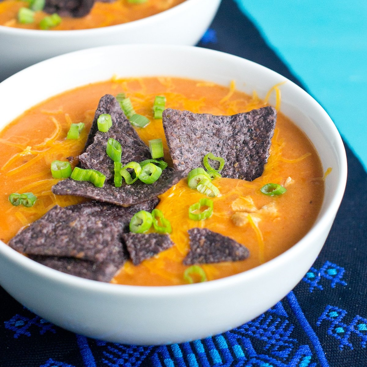 A white soup bowl with orange, cheesy Chicken Enchilada Soup, garnished with purple corn chips and green onion, set on a blue woven mat with a blue background.