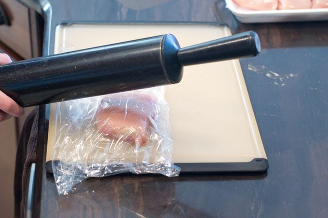 Chicken breast in plastic wrap being pounded by a black rolling pin