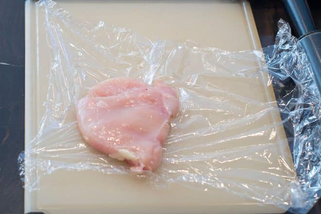 A halved chicken breast that has been pounded to an even thickness, on a piece of plastic wrap on a cutting board.
