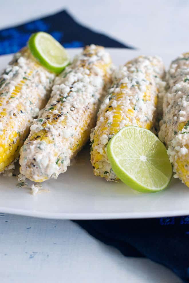 Grilled corn on the cob, Mexican elote style, with seasoning and cojita cheese coated on, on a white plate with lime slices.