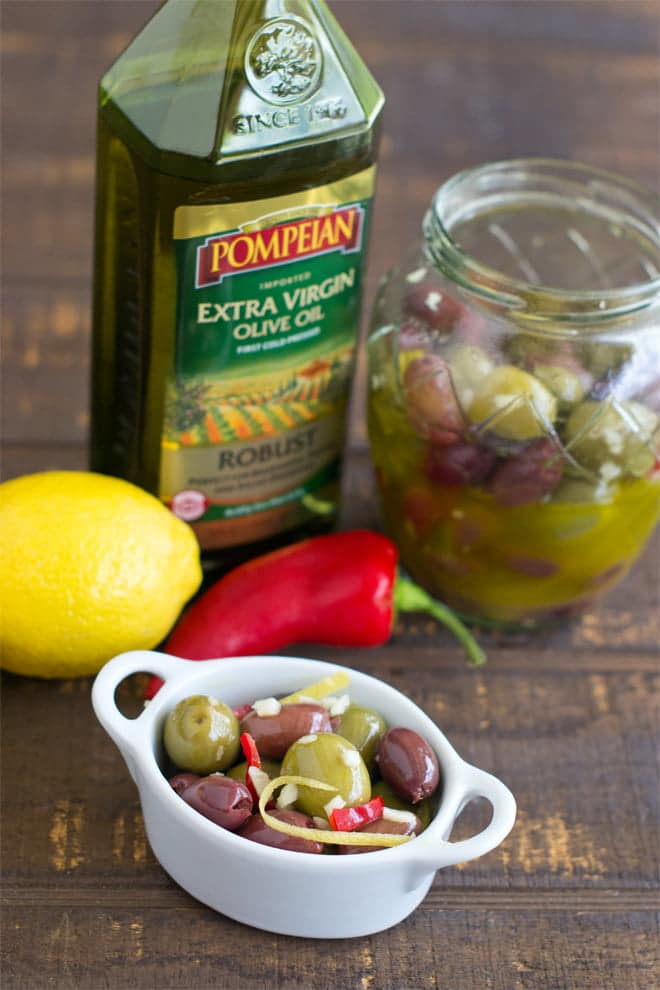 Small white dish with marinated olives, jar of olives, bottle of olive oil in background.
