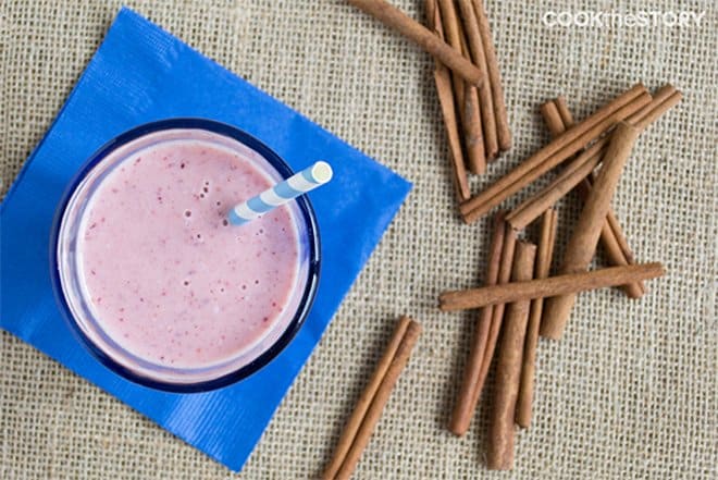 Overhead shot of cranberry smoothie in glass with straw and cinnamon sticks to the side.