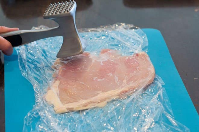 Chicken breast in plastic wrap being pounded flat with meat tenderizer.