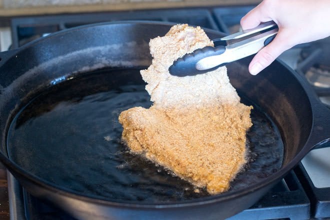 Breaded Chicken Cutlet being lowered with tongs into pan with hot oil 