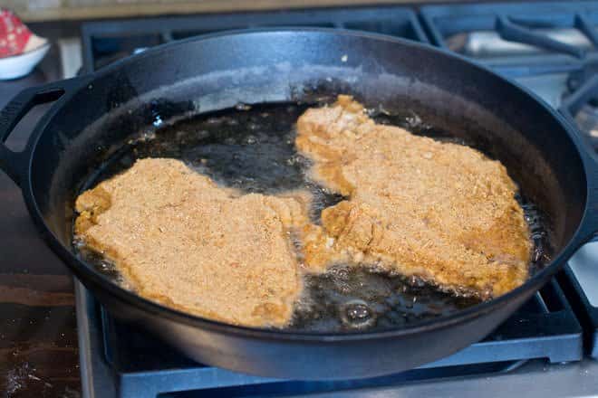 Two chicken cutlets cooking in cast iron pan with oil.