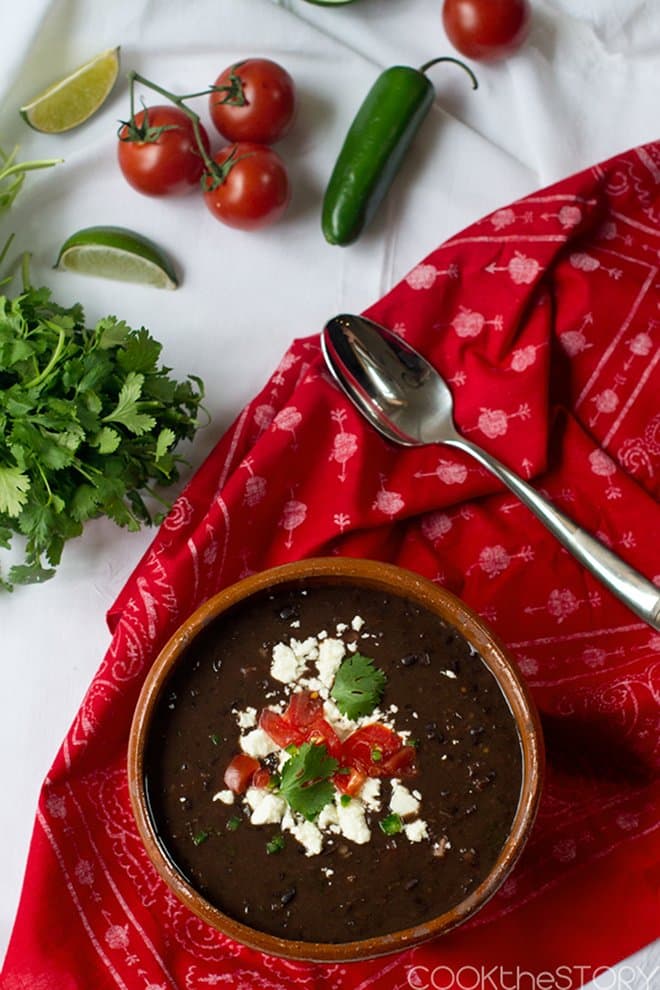 Black bean soup in a bowl topped with queso fresco, tomatoes, and parsley. On a red patterned cloth, with spoon and various fresh ingredients surrounding.