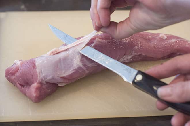 Raw pork tenderloin with the silverskin being sliced off with a knife.