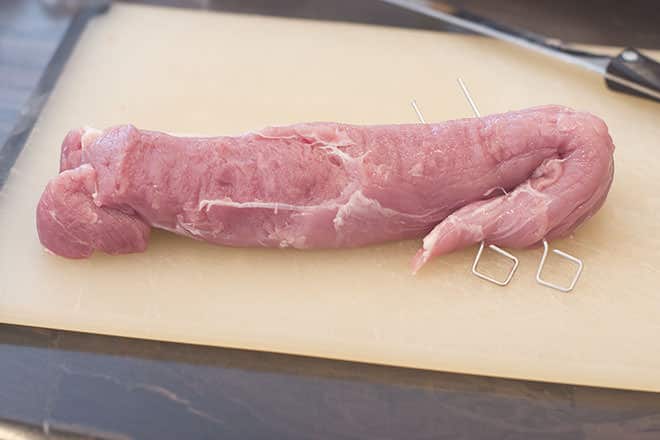 Raw pork tenderloin on a cutting board with the thinner side folded in and fastened with two metal skewers.
