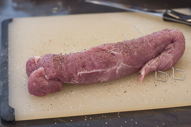 Raw pork tenderloin on cutting board that's been sprinkled with pepper and seasonings.