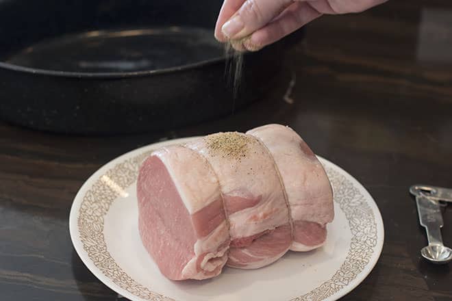 Raw pork loin on white plate with brown flowered edge. A hand is sprinkling seasoning onto the roast from above.