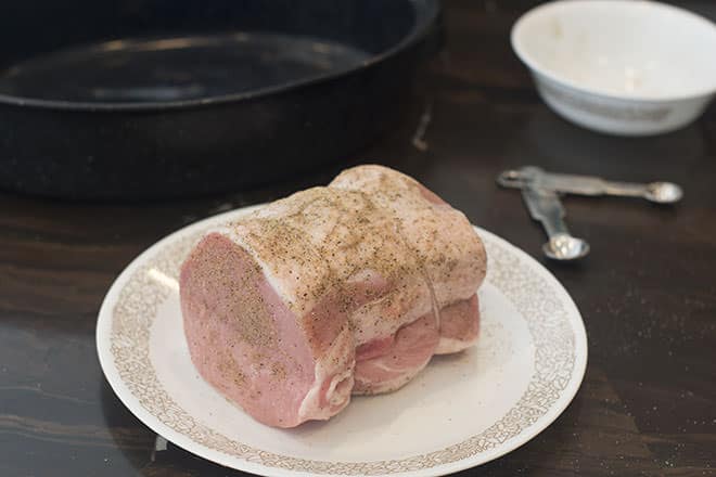 Raw pork loin on white plate with brown flowered edge. It has seasoning on it and there are spoons and a bowl in the background.