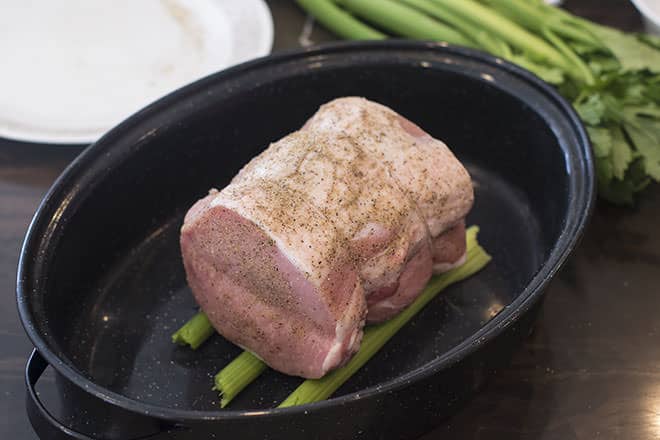 Raw pork loin in a roasting pan. The roast is seasoned and it is resting on three ribs of celery that are in the roasting pan. There is more celery and a plate behind the roasting pan.