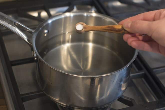 Salt on a small wooden spoon being added to water in saucepan.