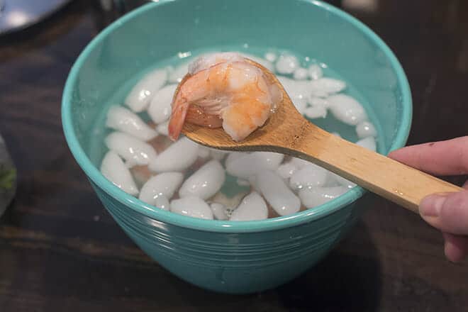 Cooked shrimp in an ice bath in a light blue bowl. A wooden spoon is being held above with a couple shrimp on it.