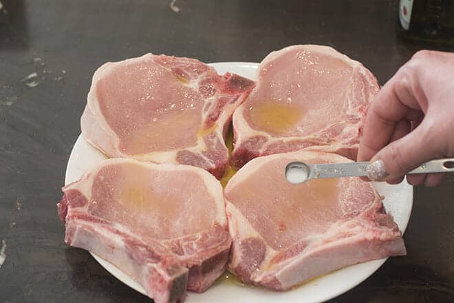 Raw, oiled pork chops being sprinkled with fine salt.
