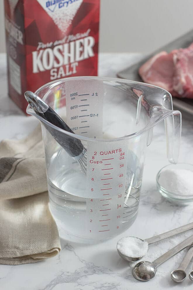 Plastic measuring cup with brine and whisk, measuring spoons with salt in foreground, box of kosher salt in background.
