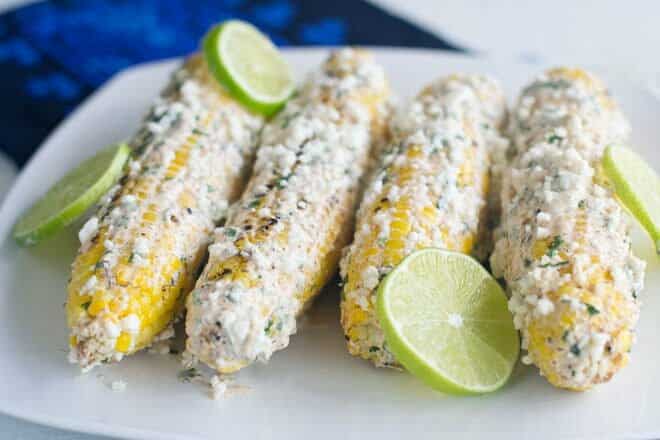 Elote corn on the cob on a white plate with lime slices.