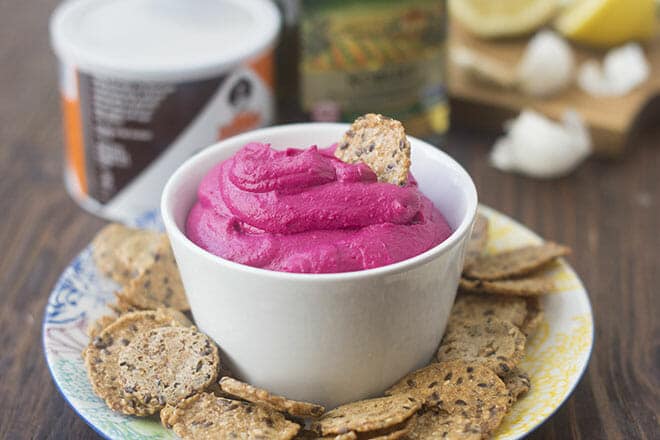 Beet hummus in white bowl, surrounded by seeded crackers.