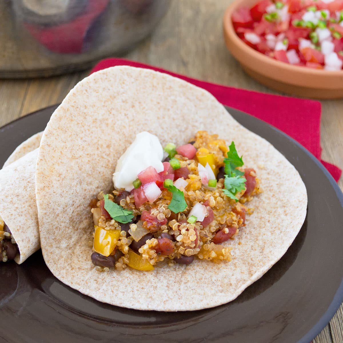 A flour tortilla on a brown plate filled with a mixture of quinoa, bell peppers, cheddar cheese melted, and cilantro, all topped with sour cream. Pico de gallo and a skillet with more quinoa mixture are in the background.