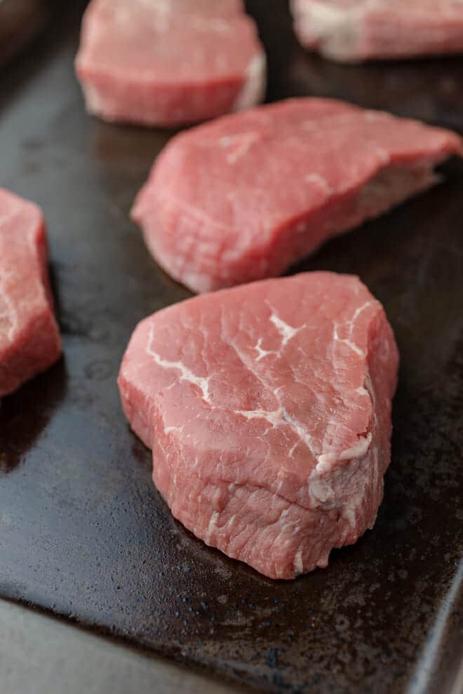 Several raw steaks sitting seperated on a baking sheet.