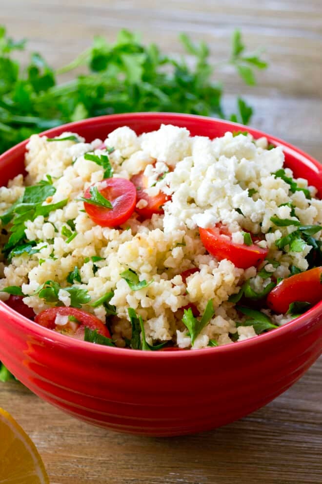A red bowl full of cooked millet topped with tomatoes, feta, and fresh parsley.