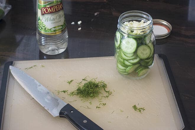 Glass jar with cucumber slices and minced garlic, fresh dill chopped on cutting board.