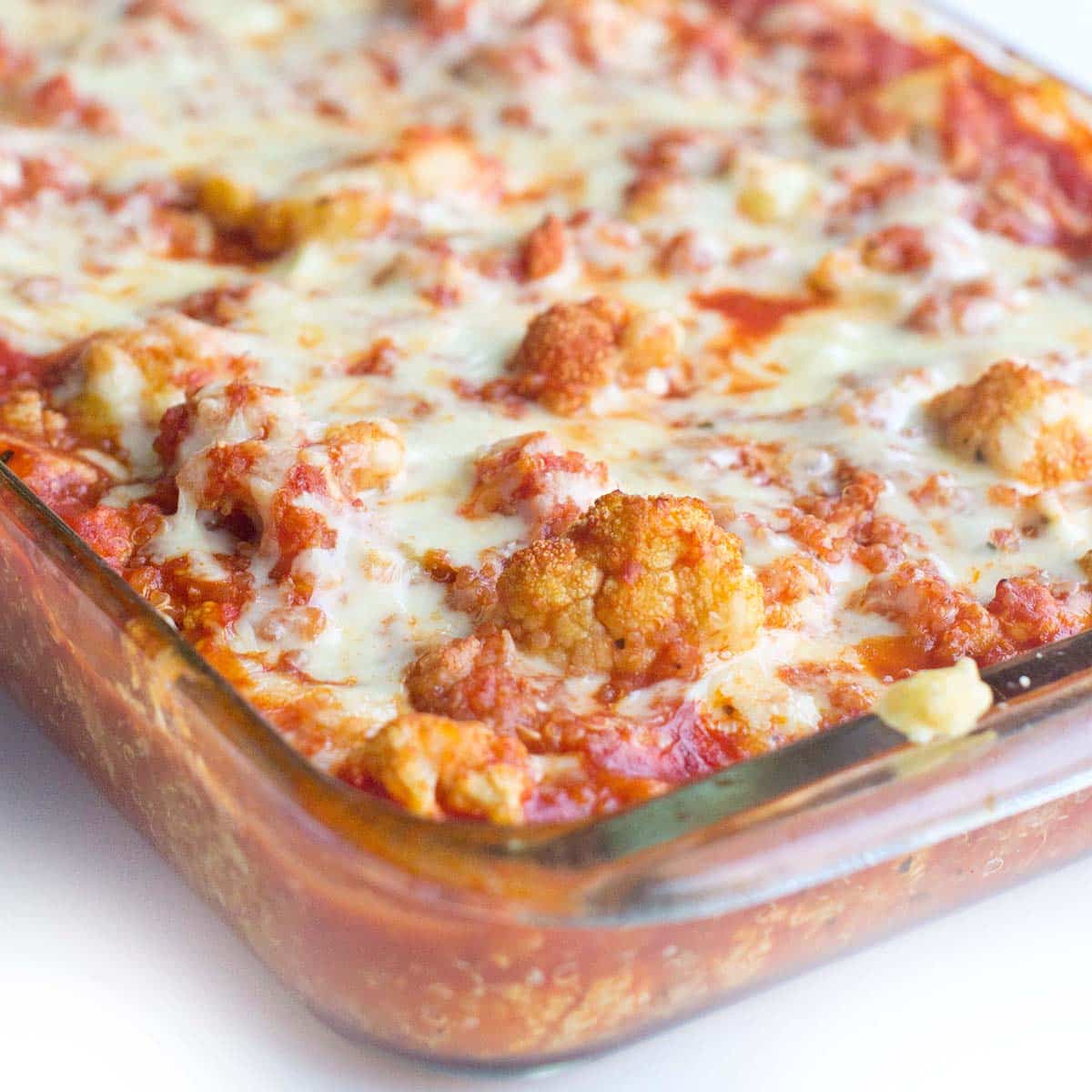 A 13x9 inch glass casserole dish filled with a mixture of cooked quinoa, cauliflower, and tomato sauce, topped with cheese. The background is all white.