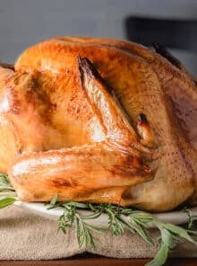 A roasted turkey that has crispy skin and a deep brown color, on a large platter surrounded by fresh sage leaves.