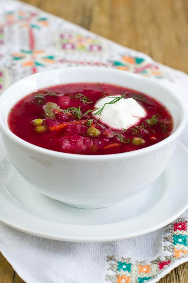 A white bowl on a white saucer with a cross-stitched linen underneath. The bowl is full of bright red beet boarscht, garnished with sour cream and dill.