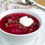 A white bowl on a white saucer with a cross-stitched linen underneath. The bowl is full of bright red beet borscht, garnished with sour cream and dill. Spoon in the background.