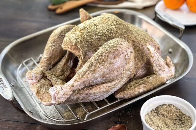 An uncooked turkey in a roasting pan covered in a dry brine mixture of salt and dried herbs.