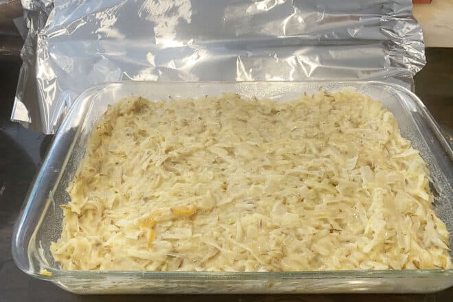 Shredded potatoes, sauce, and cheese in a 9x13 inch baking dish ready to go into the oven. A sheet of foil is next to it, ready to go on top.