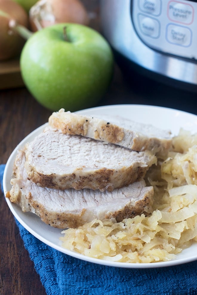 Slices of pork on top of sauerkraut on a white plate, instant pot and apple in background.