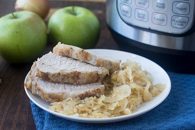 Sliced pork and sauerkraut on a white plate in front of apples and an Instant Pot.