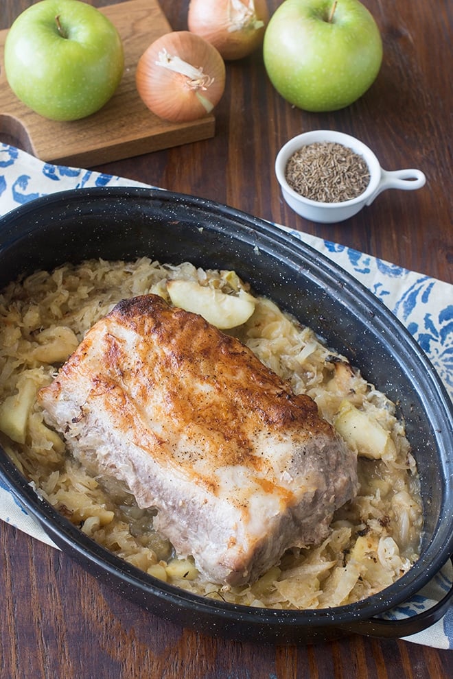 Pork and Sauerkraut in a roasting dish with apples and onions in background.
