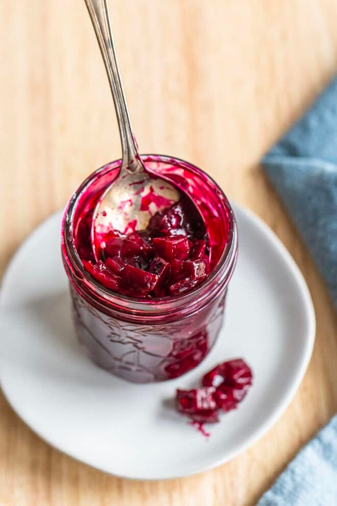 A jam jar full of scarlet beet relish on a white plate with a spoon in the jar.