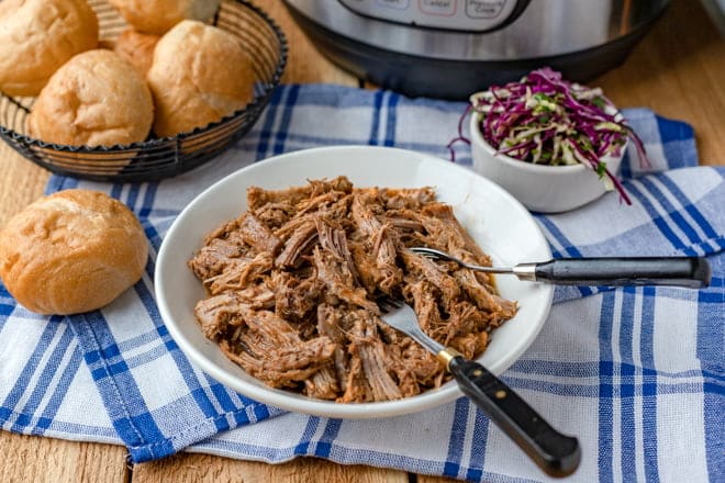 Pulled pork in a white bowl, with cabbage and rolls in background.